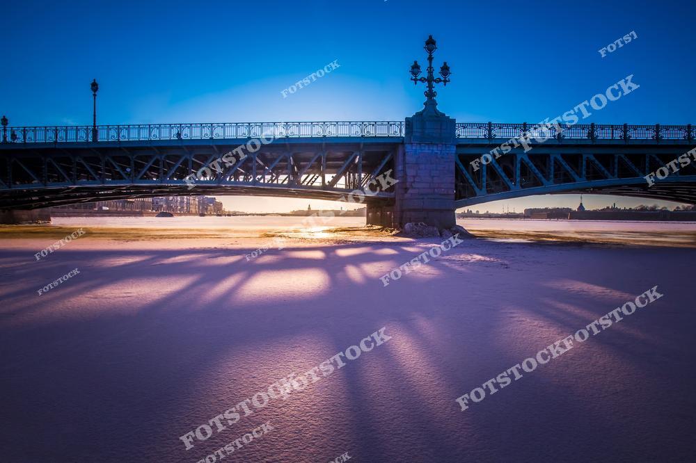 На фотографии изображён Троицкий мост — один из самых известных мостов Санкт-Петербурга, соединяющий Петроградскую сторону с центральной частью города. Под мостом видна замёрзшая поверхность Невы, освещённая лучами зимнего солнца. Мост украшен коваными фонарями и декоративными элементами в историческом стиле. На горизонте просматриваются очертания Петропавловской крепости и других знаковых зданий. Атмосфера утреннего покоя подчёркивает красоту петербургской зимы.